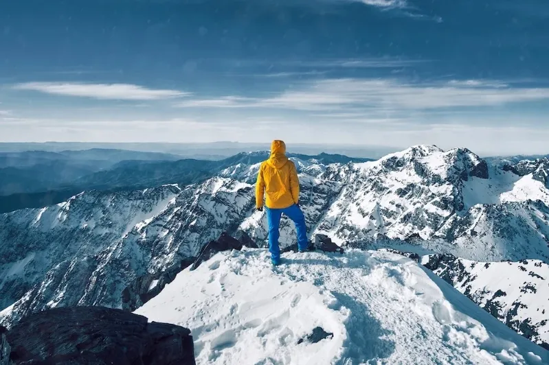 Man in a yellow jacket standing triumphantly on a mountain peak in the Atlas Mountains of Morocco.