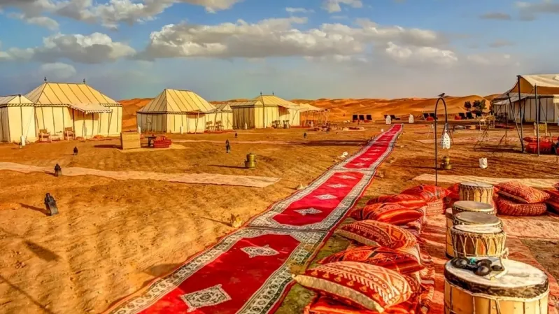 A luxury camp in the Moroccan desert featuring tents set on a vibrant carpet against a sandy landscape.