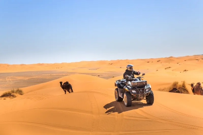 A man riding an ATV through the desert near a luxury camp in Morocco, surrounded by sandy dunes and clear blue skies.
