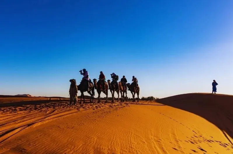 People riding camels in the desert at sunset, showcasing a Marrakech tours experience. 